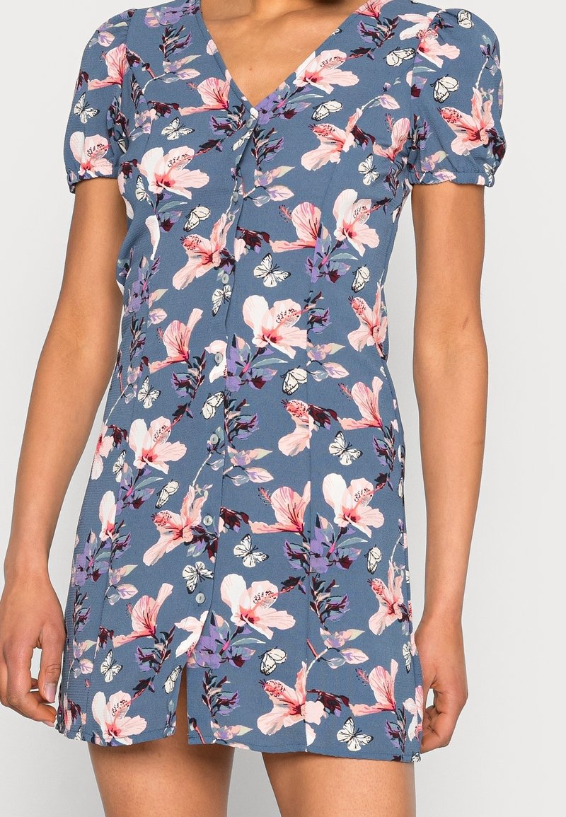 Mini-robe florale en tissu bleu avec des fleurs roses et blanches ainsi que des papillons. Encolure en V avec manches courtes bouffantes et détails de boutons.