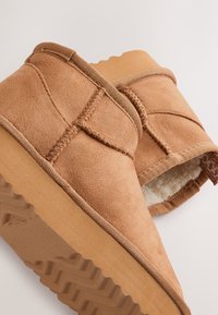 Hellbraune Wildlederschlappen mit weichem Futter, ausgestattet mit einer Gummisohle, gesteppten Details und offenem Rücken für einfaches An- und Ausziehen.