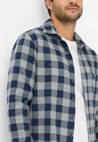 Blauw en grijs geruit overhemd van stof met een gestructureerd gevoel. Heeft een knoopsluiting aan de voorkant, een kraag en twee borstzakken.