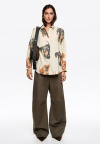 Koszula z długim rękawem w kolorze kremowym z dużymi, nadrukowanymi twarzami psów; połączona z szerokimi, oliwkowo-zielonymi spodniami. Posiada zapinany na guziki przód i czarną torbę na ramię.