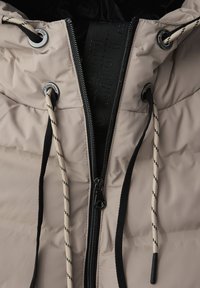 Pufjakke i lys beige, med lynlås lukning, snoretræk og en struktureret overflade. Inkluderer kontrasterende sorte og beige accenter.