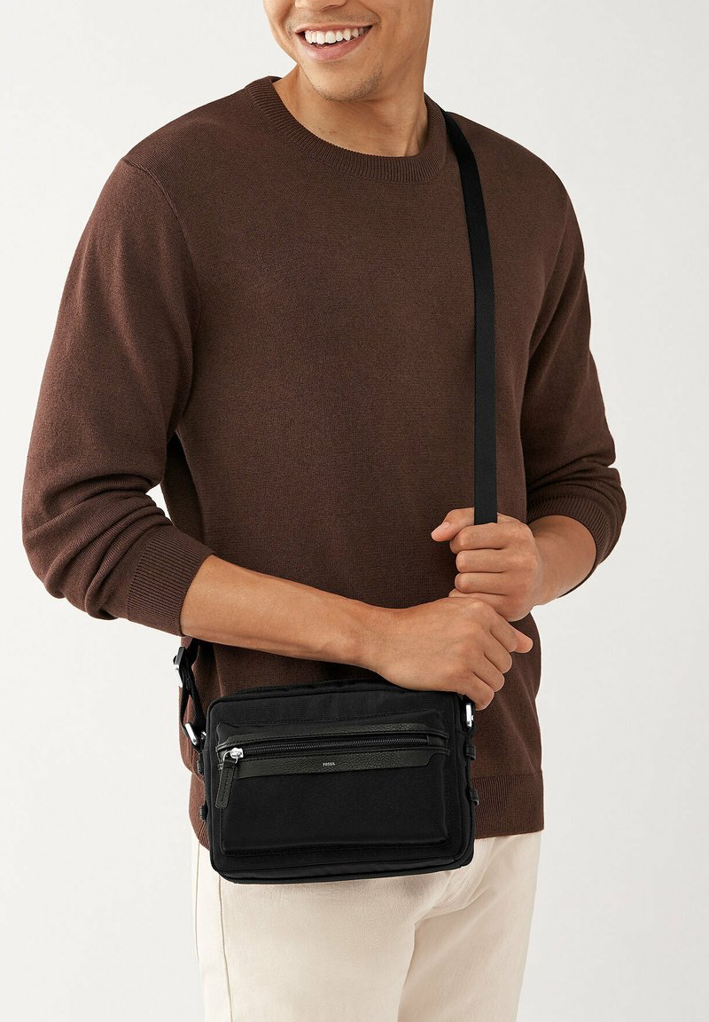 Borsa a tracolla nera realizzata in tessuto resistente, dotata di diverse tasche con cerniera, una superficie texturizzata e una tracolla regolabile.