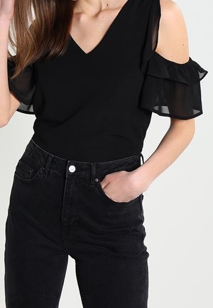Kvinde iført en sort cold-shoulder top med flæseærmer og sorte jeans med høj talje, hånd i lommen.