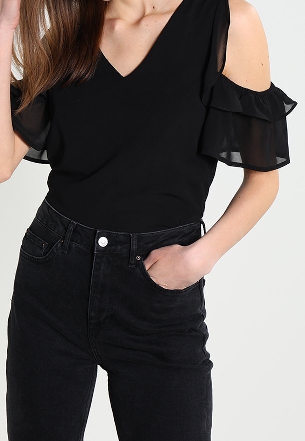 Femme portant un top noir épaules dénudées avec des manches à volants et un jean noir taille haute, main dans la poche.