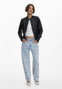 Czarna skórzana kurtka ze stójką, zestawiona z jasnoniebieskimi dżinsami o szerokich nogawkach, ozdobionymi guzikami wzdłuż boku, oraz białymi trampkami.