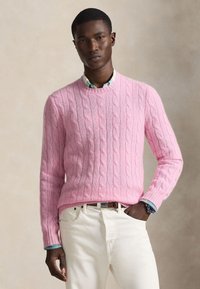 Homme portant un pull en maille torsadée rose par-dessus une chemise à motifs, pantalon blanc, main dans la poche, debout devant un fond neutre.
