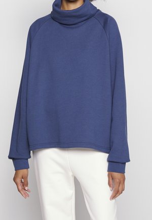 Osoba ubrana w luźną, niebieską bluzę z golfem i białe spodnie, stojąca na tle jednolitego, jasnego tła.