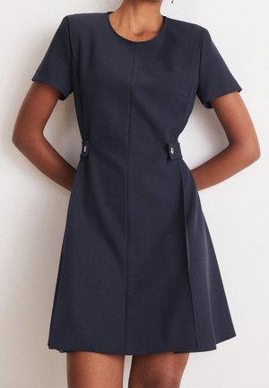 Navy sininen lyhythihainen mekko, jossa on pyöreä kaula-aukko, muotoiltu leikku, sivutaskut ja koristeelliset napit vyötäröllä. Silkkinen tekstuuri.