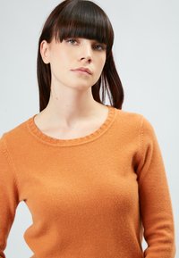 Suéter naranja con un amplio cuello redondo acanalado. Hecho de una tela suave y texturizada, presenta mangas largas y una silueta ajustada.