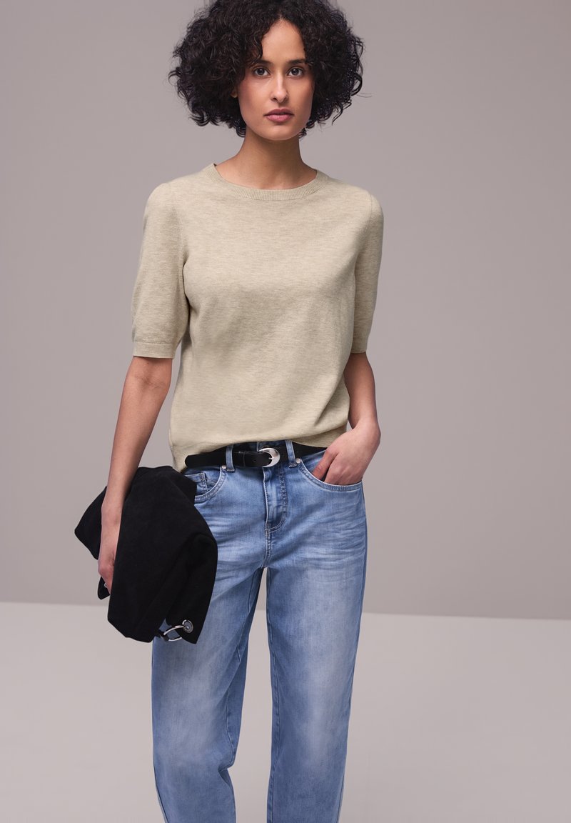 Donna con capelli ricci che indossa un maglione beige a maniche corte e jeans blu, tiene una giacca nera nella mano sinistra su sfondo semplice.