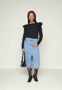 Jovem mulher vestindo uma blusa preta com babados de mangas longas, uma saia midi de denim azul claro com fenda frontal, botas pretas bordadas, e segurando uma bolsa preta.