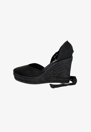 Zapato espadrille negro con cuña, puntera cerrada, tacón tejido y lazos para atar en el tobillo sobre un fondo blanco.