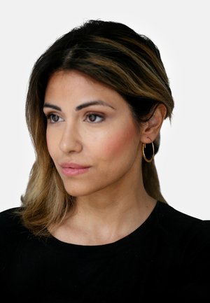 Eine goldene Creolen-Ohringe, die von einer Frau getragen wird, mit einer glatten, glänzenden Oberfläche. Ihr Haar ist braun mit helleren Strähnen und in sanften Wellen gestylt.