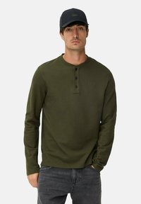Olijfgroene longsleeve shirt met een ronde hals, drie zwarte knopen aan de voorkant en een subtiele textuur. Gecombineerd met grijze jeans.