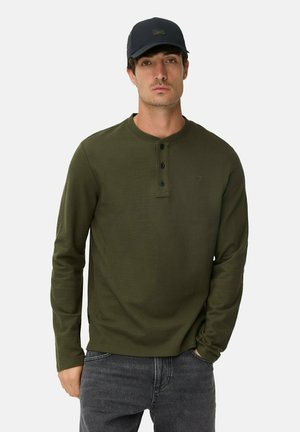 Olivenfarvet skjorte med lange ærmer og rund halsudskæring, tre sorte knapper foran og en subtil tekstur. Kombinationen er med grå jeans.