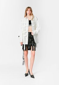 Chaqueta blanca de gran tamaño con diseño de cuadrícula, top corto negro, falda plisada negra con tirantes y zapatos de tacón negro.