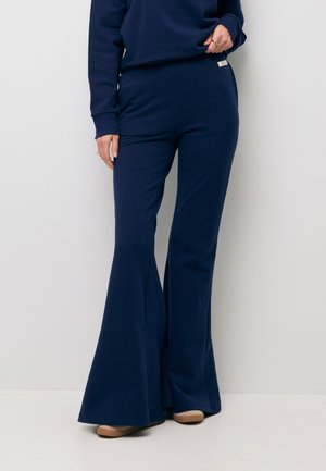 Persoon draagt marineblauwe wijde broek en bijpassende trui, beige schoenen, staand tegen een lichtgrijze muur.