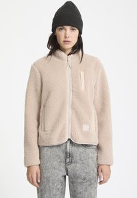Lichtbeige fleecejack met een ritssluiting aan de voorkant, twee zijzakken en een vierkant patchdetail. Gecombineerd met grijze jeans en een zwarte beanie.