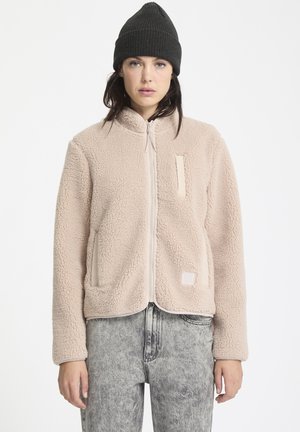 Lichtbeige fleecejack met een ritssluiting aan de voorkant, twee zijzakken en een vierkant patchdetail. Gecombineerd met grijze jeans en een zwarte beanie.