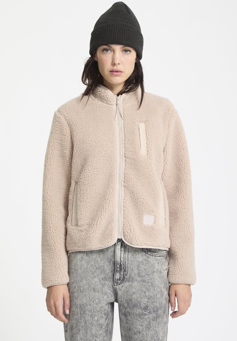 Lichtbeige fleecejack met een ritssluiting aan de voorkant, twee zijzakken en een vierkant patchdetail. Gecombineerd met grijze jeans en een zwarte beanie.
