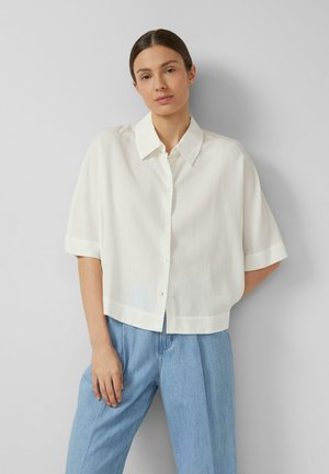 Frau mit dunklen Haaren trägt ein locker sitzendes weißes kurzärmeliges Hemd mit Knöpfen und hellblaue, hoch taillierte Jeans, steht vor einer grauen Wand.