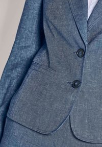 Close-up van een blauwe denim blazer met twee donkere knopen en een klepzak aan de onderzijde, gedragen over een wit shirt.
