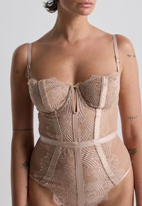Body in pizzo color nude con spalline in satin e sottoseno. Presenta pizzo floreale, una scollatura a chiave e una silhouette aderente.