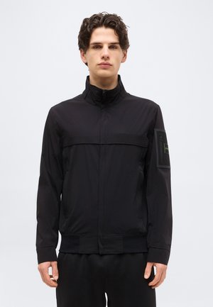 Jeune homme aux cheveux bouclés portant une veste noire à fermeture éclair avec un col montant et une poche patch sur la manche gauche.