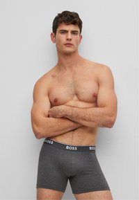 Graue Boxershorts mit einem schwarzen Bündchen, das mit dem "BOSS"-Logo versehen ist. Der Stoff wirkt glatt, passgenau und flexibel. Das Model präsentiert sich mit verschränkten Armen.
