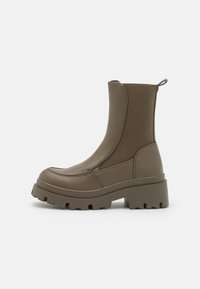 Botas de tobillo verde oliva hechas de un material sintético liso con una suela de goma gruesa, que cuentan con paneles elásticos a los lados y una lengüeta.