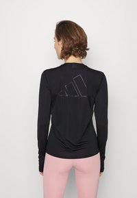 Schwarz-langärmliges Sportshirt mit glatter Textur. Auf der Rückseite ist ein tonal Adidas-Logo zu sehen. Kombiniert mit pinken Leggings.