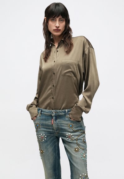 Olivovo zelená hodvábna košeľa s vreckom, kombinovaná s modrými džínsami zdobenými dekoratívnymi nitmi a farebnými ozdobami pozdĺž strán.