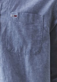 Camisa azul de botones con un bolsillo en el pecho que presenta un pequeño logo. El material parece texturizado con un acabado suave y ligeramente arrugado.