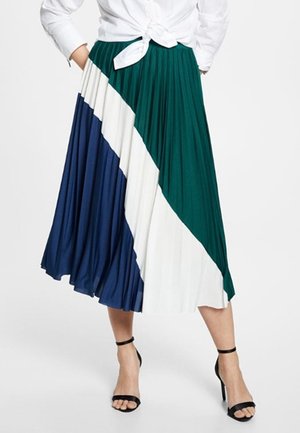 Femme portant une jupe midi plissée avec des blocs de couleurs vert, blanc et marine, associée à une chemise blanche nouée et des talons noirs à bride cheville.