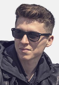 Black sunglasses with dark lenses, a sleek rectangular frame, worn by a person in a black hoodie and leather jacket. Short, styled hair.