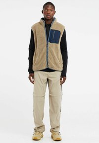 Beige fleecetype vest met een marineblauwe zak, voorzien van een hoge kraag en een ritssluiting. Draag het over een zwart shirt met lange mouwen en combineer het met lichte broek.