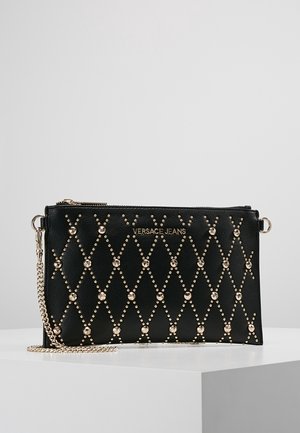 Clutch negra de Versace Jeans con patrón de diamantes tachonado en tono dorado y una correa de cadena dorada sobre superficie blanca.