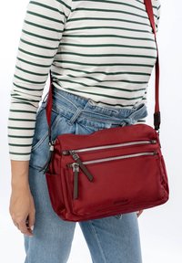 Nainen, jolla on siniset farkut ja vihreäraitaiseen pitkähihnaiseen paitaan sekä punainen olkalaukku, jossa on kaksi etuosa vetoketjutaskua.