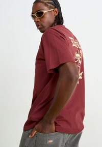 T-shirt bordeaux avec un motif graphique dans le dos, en tissu coton, à manches courtes, associé avec un pantalon en denim gris et des lunettes de soleil claires.