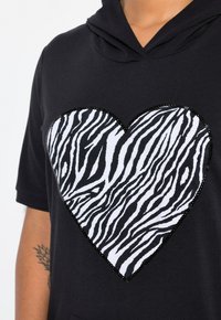 Felpa nera con una grande toppa a forma di cuore che presenta un motivo a strisce zebrate bianche e nere, bordato da paillettes nere. Tessuto morbido.