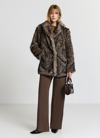 Cappotto in pelliccia sintetica marrone con un ampio colletto, abbinato a pantaloni marroni a trama. Tiene una piccola borsa scura e indossa scarpe nere a punta.