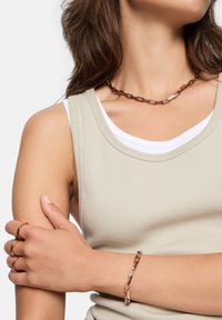 Halsband och armband i roséguld, med länkade ovala former. Modellen bär en ljusgrå ribbad linne över en vit skjorta.