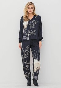 Woman wearing black floral print loose-fit long-sleeve top and matching pants with black ankle boots, standing against a plain light background.