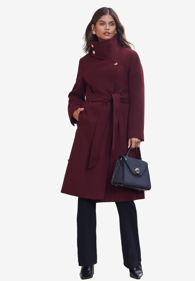 Donna che indossa un cappotto bordeaux con cintura e collo alto, pantaloni neri, stivali neri, orecchini dorati, che tiene una borsa nera con chiusura metallica.