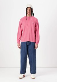 Long-sleeve, pink shirt with a small white logo, paired with loose, dark blue jeans and cream-colored sneakers, topped with a light cap.