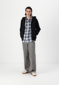 Schwarze Zip-Up Kapuzenjacke, blau-weiß karierte Hemd, grau gestreifte weite Hose und weiße Sneaker mit braunen Sohlen. Einfaches lässiges Outfit.