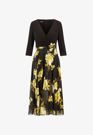 Schwarzes Kleid mit tailliertem Oberteil, V-Ausschnitt und 3/4-Ärmeln. Der fließende Rock weist ein gelbes Blumenmuster und ein Stoffband mit einem Blütendetail auf.