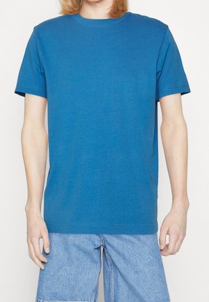 Personne portant un T-shirt bleu uni à manches courtes et un short en denim bleu clair, debout avec les bras détendus le long du corps.