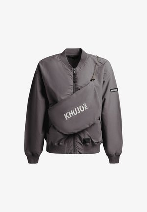 Chaqueta bomber gris con cremallera, cuello y puños acanalados, combinada con un bolso bandolera a juego que lleva el logo "KHUJO 2001" en blanco.