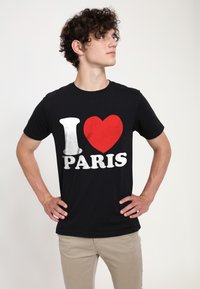 Mladý muž s kudrnatými vlasy má na sobě černé tričko s nápisem "I ♥ PARIS" v bílé a červené barvě, stojí s rukama na bocích a dívá se stranou.